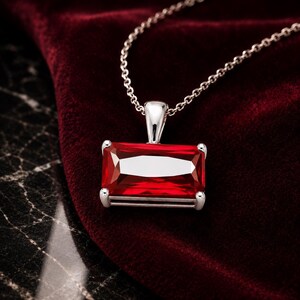 May include: A rectangular, faceted red gemstone pendant set in a silver-toned metal frame, suspended from a delicate silver-toned chain. The vibrant red stone features sharp, clean edges and a polished finish, reflecting light across its surface.