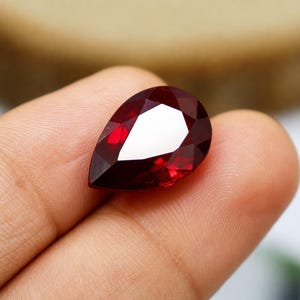 Può includere: Una gemma sfaccettata a forma di pera di un colore rosso intenso e vibrante. La pietra presenta una superficie lucida con molteplici sfaccettature triangolari e a forma di aquilone che riflettono la luce in modo brillante.