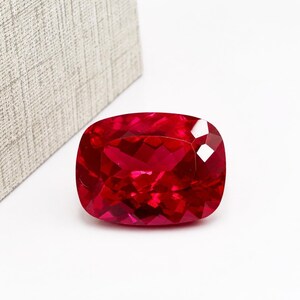Pode incluir: Uma pedra preciosa de cor vermelha profunda e vibrante com um corte almofada e uma superfície facetada que reflete a luz. A pedra tem uma forma retangular com cantos arredondados e um acabamento polido, exibindo uma cor rica e saturada.