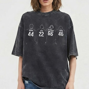 May include: A dark grey t-shirt with a graphic of four figures, each with a name and number: Graham 44, Logan 22, Di Laurentis 66, and Tucker 46. The tee has a vintage, washed look and a crew neck.