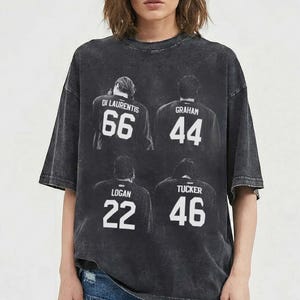 Peut inclure: T-shirt gris foncé surdimensionné avec un aspect vintage délavé. Le t-shirt présente quatre noms et numéros de joueurs : Di Laurentis 66, Graham 44, Logan 22 et Tucker 46. Le design est en blanc.