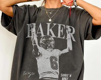 Puede incluir: Una camiseta gris carbón de manga corta con un gráfico blanco desgastado de un jugador de fútbol americano con el número 6. El texto BAKER aparece sobre el jugador, junto a SHAKE 'N BAKE y una firma en la parte inferior.