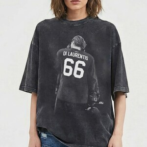 May include: A charcoal gray t-shirt featuring a graphic of a person wearing a jersey with the name "DI LAURENTIS" and the number "66" in white. The shirt has a vintage, washed appearance.