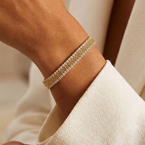 May include: A gold-toned bracelet featuring a central woven mesh band flanked by two rows of small, white, spherical beads. The bracelet has a structured, multi-layered design with a metallic finish and a consistent, repeating pattern throughout its length.