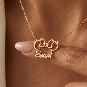 May include: A gold-colored necklace featuring a bow and mouse ears design, with the name "Sarah" inscribed below. The pendant hangs from a delicate chain. The necklace is displayed against a white background.