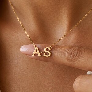 May include: A gold-toned necklace featuring a delicate chain and a pendant consisting of the capital letters A and S, separated by a small, open-work heart symbol. The pendant is centered on the chain, which rests against the collarbone area.