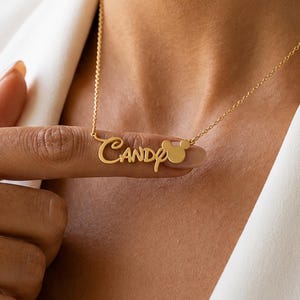 May include: A gold-toned metal necklace featuring a delicate chain and a custom nameplate pendant that displays the name Candy in a cursive script font, followed by a small, solid circular icon resembling a mouse head silhouette.