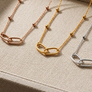 Può includere: Tre delicate collane nei toni dell'oro rosa, oro giallo e argento. Ogni collana presenta una catenina sottile con piccole perle rotonde distanziate uniformemente e un punto focale centrale composto da due maglie ovali intrecciate.