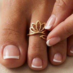 Puede incluir: Un anillo para el dedo del pie de metal dorado con un diseño de flor de loto estilizada. El anillo tiene una banda abierta que se envuelve alrededor del dedo, mostrando un motivo de pétalos simétricos con un acabado pulido que refleja la luz.