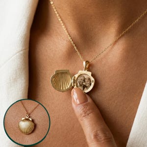 Op de afbeelding: Een goudkleurige, schelpvormige medaillonhanger aan een fijne ketting. Het medaillon is geopend afgebeeld en toont een kleine zwart-witfoto van twee personen, terwijl een cirkelvormig inzetstuk het medaillon in gesloten toestand laat zien.