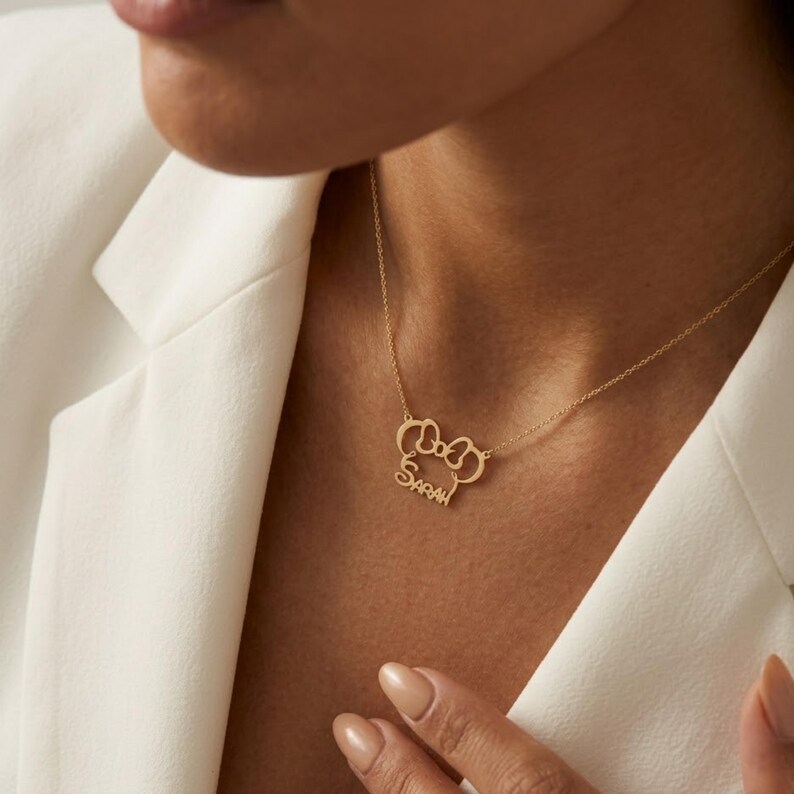 May include: A gold-colored necklace with a bow-shaped pendant and the name "Sarah" in a delicate script. The necklace is worn against a white blazer, highlighting the jewelry's design.
