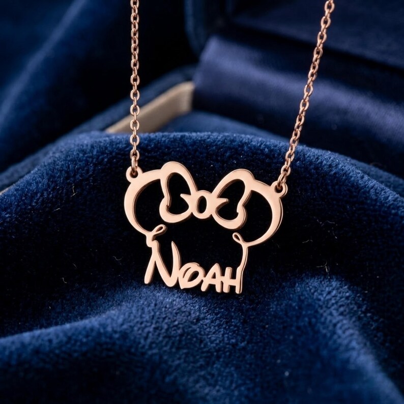 May include: Rose gold necklace featuring the name "Noah" in a stylized font, with a bow-shaped design above the name. The necklace is displayed against a dark blue velvet background.