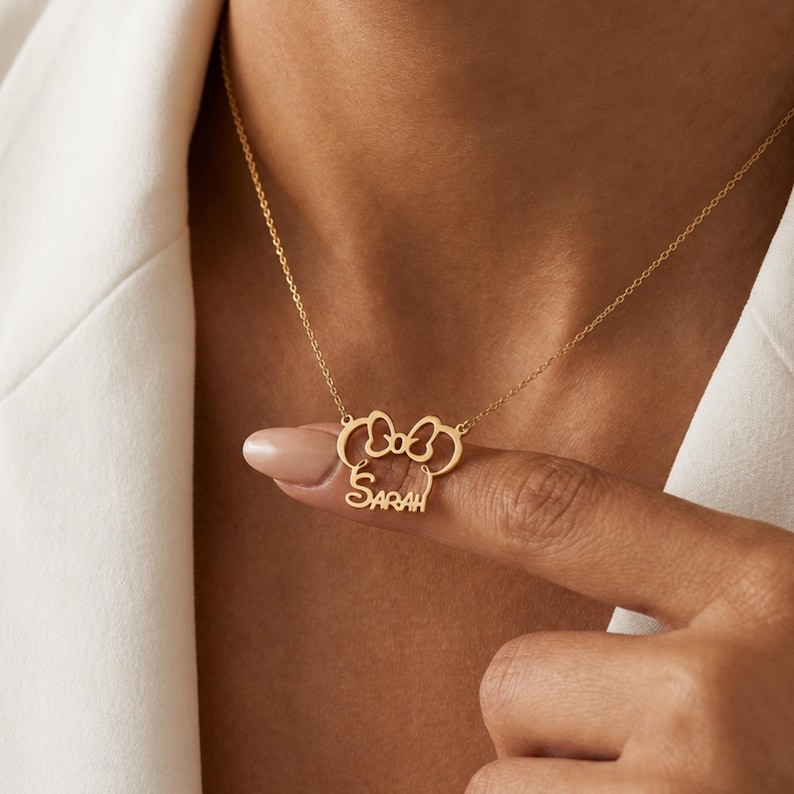 May include: A gold-colored necklace featuring a bow and mouse ears design, with the name "Sarah" inscribed below. The pendant hangs from a delicate chain. The necklace is displayed against a white background.