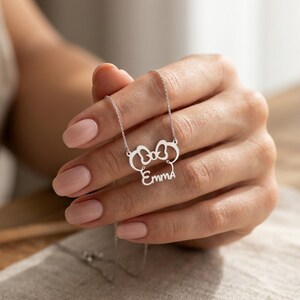May include: A silver-colored necklace featuring a bow-shaped pendant with the name "Emma" in a cursive font. The necklace is held by a person with manicured nails.
