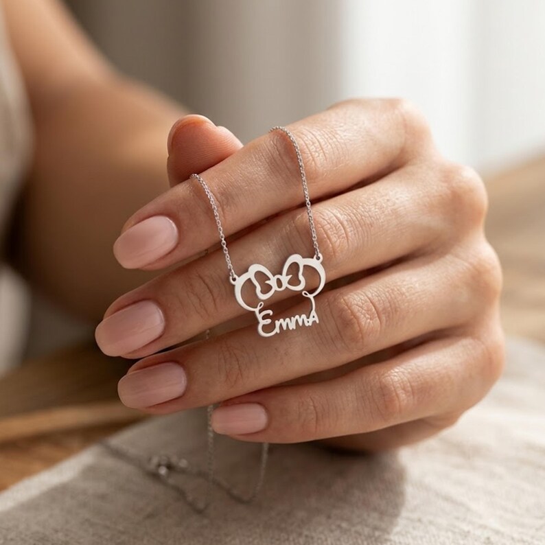 May include: A silver-colored necklace featuring a bow-shaped pendant with the name "Emma" in a cursive font. The necklace is held by a person with manicured nails.
