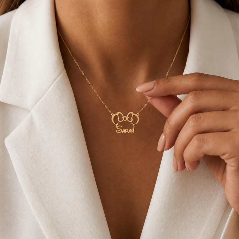 May include: Gold-colored necklace with a bow-shaped pendant and the name "Sarah" in cursive script. The necklace is worn against a white blazer.