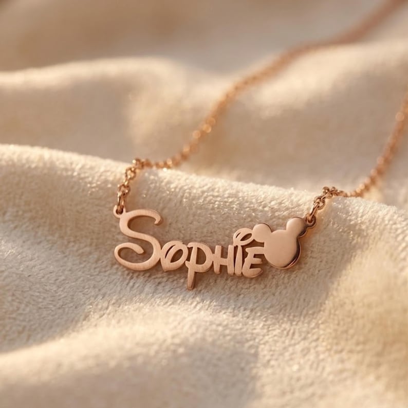 May include: A rose gold-toned necklace featuring a delicate chain and a custom nameplate pendant that displays the name Sophie in a cursive script font, with a small, solid mouse-ear silhouette icon attached to the end of the name.