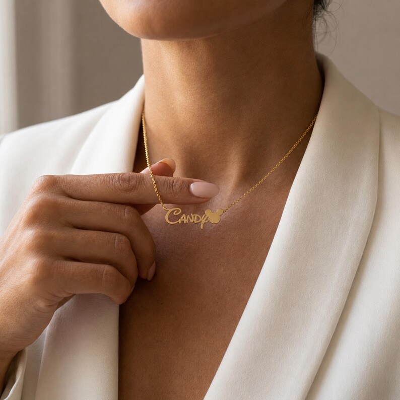 May include: A gold-toned metal necklace featuring a delicate chain and a personalised nameplate pendant. The pendant displays the name Candy in a cursive script font, with a small circular icon resembling a mouse head silhouette attached.