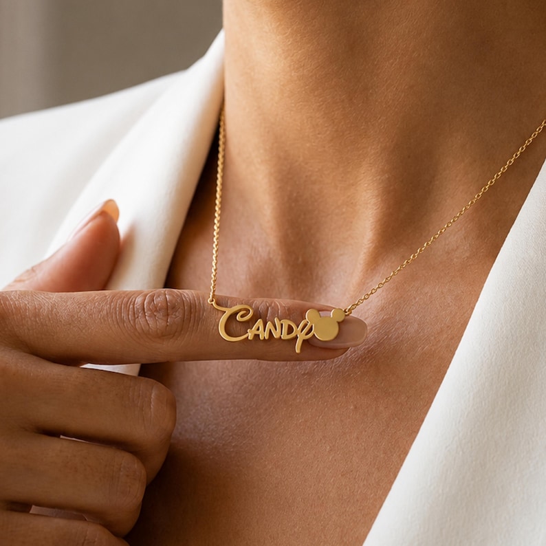 May include: A gold-toned metal necklace featuring a delicate chain and a custom nameplate pendant that displays the name Candy in a cursive script font, followed by a small, solid circular icon resembling a mouse head silhouette.