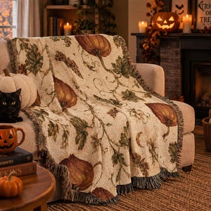 May include: A woven throw blanket featuring a repeating pattern of orange pumpkins, green leaves, and vines on a cream background with fringed edges, draped over a sofa alongside a black cat, a white pumpkin, and a jack-o'-lantern mug.