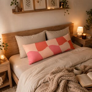 Pode incluir: Uma almofada retangular comprida com um padrão de xadrez ondulado em tons de coral, rosa e creme. A almofada está posicionada horizontalmente sobre uma cama com roupa de cama clara, apoiada contra duas almofadas bege lisas.