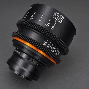 May include: A black camera lens featuring a prominent orange ring, geared focus and aperture control rings, and white printed markings including 58, T2.1, and M42-NEX. The lens has a cylindrical body with textured grip sections for manual adjustments.