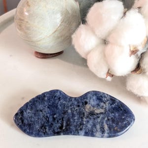 Könnte beinhalten: Ein poliertes, flügelförmiges Gua-Sha-Werkzeug aus tiefblauem Sodalith-Stein mit gesprenkelten schwarzen und weißen Einschlüssen. Die glatte, geschwungene Oberfläche weist eine ausgeprägte mittlere Vertiefung auf, die die natürlichen Mineralmuster zeigt.