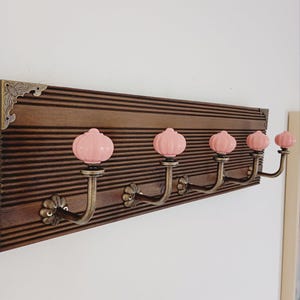 Peut inclure: Un porte-manteau mural composé d'une planche en bois brun foncé à rainures horizontales avec des coins décoratifs en métal. Il comporte cinq crochets en métal couleur laiton, chacun surmonté d'un bouton en céramique rose pâle.