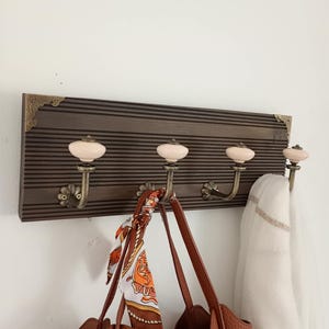 Peut inclure: Un porte-manteau mural composé d'une planche en bois brun foncé à rainures horizontales avec des coins décoratifs en laiton. Il comporte quatre crochets en métal couleur laiton, chacun surmonté d'un bouton en céramique crème lisse.