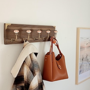 Peut inclure: Un porte-manteau en bois avec cinq crochets décoratifs, chacun orné d'un bouton en céramique à motifs floraux. Un sac à main en cuir marron et une veste à carreaux sont suspendus aux crochets. Une image encadrée est accrochée au mur.
