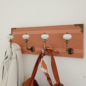 Peut inclure: Un porte-manteau mural rectangulaire avec une base en bois strié de couleur saumon et quatre crochets en laiton. Chaque crochet est surmonté d'un bouton en céramique blanche ronde et présente des coins décoratifs en laiton sur la base.
