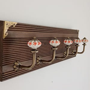 Peut inclure: Un porte-manteau mural en bois avec quatre crochets. Le bois brun foncé présente un motif rainuré. Les crochets sont de couleur bronze avec des boutons en céramique florale blanche et rouge. Un coin décoratif en laiton est visible.