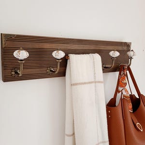 Op de afbeelding: Een rechthoekig wandrek van donkerbruin hout met vier metalen haken voorzien van witte keramische knoppen met gouden bloemmotieven. Het rek heeft decoratieve metalen hoekaccenten. Een witte handdoek met ruitpatroon en een bruine leren handtas hangen aan de haken.