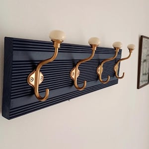 Op de afbeelding: Een wandkapstok met een donkerblauwe, rechthoekige houten basis met horizontale ribbels. Vier messingkleurige metalen haken zijn aan het bord bevestigd, elk bekroond met een gladde, ronde, crèmekleurige keramische knop.