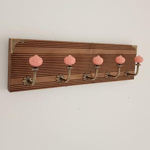 May include: A rectangular wall-mounted coat rack featuring a dark brown ribbed wooden base with decorative metal corner accents. It includes five brass-toned metal hooks, each topped with a round, ribbed, light pink ceramic knob.