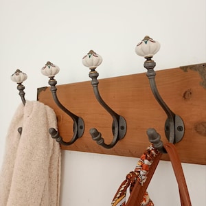 Può includere: Un appendiabiti da parete con una tavola di legno rettangolare e quattro ganci in metallo scuro. Ogni gancio è sormontato da un pomello in ceramica bianco a forma di zucca con piccoli dettagli floreali. Una sciarpa beige e una borsa marrone.