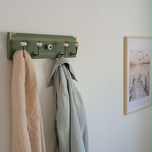 May include: A rectangular wall-mounted coat rack with a sage green ribbed wooden base and gold-toned corner accents. It features four bronze-colored metal hooks topped with small white ceramic knobs, with an additional matching knob in the center.