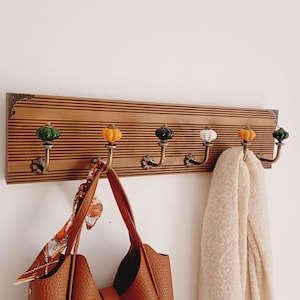 Peut inclure: Un porte-manteau mural rectangulaire en bois à rainures horizontales, équipé de six crochets en métal couleur laiton. Chaque crochet est surmonté d'un petit bouton en céramique en forme de citrouille, alternant les couleurs vert, jaune, noir et blanc.