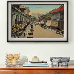 May include: A framed vintage print depicting a street scene in Escolta, Manila, featuring historic buildings, early 20th-century automobiles, and horse-drawn carriages. The print is displayed in a black frame with a white mat board.