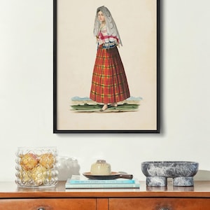 May include: A framed print featuring a historical illustration of a person wearing a white lace head covering, a pink and white striped top, and a long red plaid skirt. The artwork is displayed in a thin black frame on a light-colored wall.