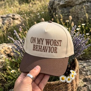 May include: A two-tone baseball cap featuring a cream-colored crown and a dark brown brim. The front of the crown displays the embroidered text ON MY WORST BEHAVIOR in dark brown thread. The cap has a structured front panel and a button top.