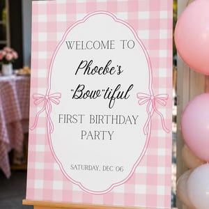 May include: A pink and white gingham welcome sign on a wooden easel. The sign has an oval frame with bows and the text "WELCOME TO Phoebe's "Bow-tiful" FIRST BIRTHDAY PARTY SATURDAY, DEC 06". Pink balloons are visible in the background.