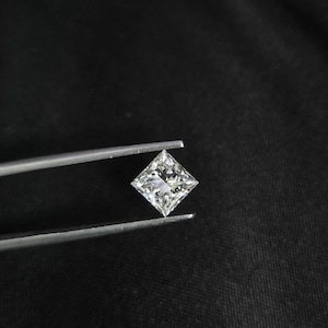 以下が含まれることがあります： シルバーカラーの金属製ピンセットで保持された、透明で無色のプリンセスカットの宝石。正方形の石には複数の三角形のファセットがあり、光を反射して暗い背景に対して輝きを放つ外観を作り出しています。