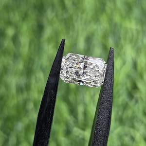 Può includere: Una gemma rettangolare incolore e trasparente con taglio radiante, tenuta da una pinzetta in metallo nero. La pietra presenta una complessa disposizione di sfaccettature triangolari e a forma di aquilone che creano un aspetto brillante.