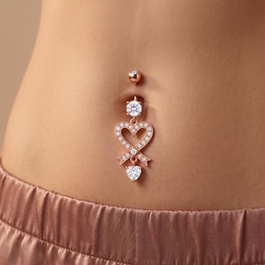 May include: A rose gold navel piercing featuring a dangling heart-shaped charm encrusted with small clear stones. The piece includes a round stone at the top and a larger heart-shaped stone at the bottom, connected by a ribbon-like heart frame.
