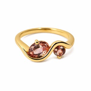 Peut inclure: Une bague en or dotée d'un anneau lisse et ondulé qui entoure deux pierres précieuses facettées. La plus grande pierre est de forme ovale et de couleur rose brunâtre, tandis que la plus petite est ronde et présente la même teinte translucide.