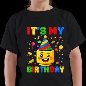 May include: A black t-shirt featuring a graphic of a yellow winking toy-style head wearing a colorful party hat. The text "IT'S MY BIRTHDAY" is printed in blocky, multi-colored letters resembling building bricks, surrounded by confetti and small bricks.