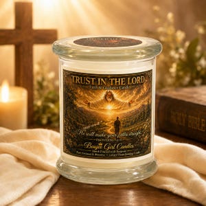 Puede incluir: Una vela grande de vidrio con tapa, con una etiqueta con el texto "TRUST IN THE LORD" y una imagen de una figura en las nubes. La vela está etiquetada como "Boujee Girl Candles" y está sobre una superficie de madera.