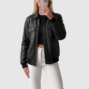 Puede incluir: Una chaqueta bomber de cuero negro con acabado texturizado, cierre de cremallera frontal, dos bolsillos en el pecho con solapas abotonadas, y puños y dobladillo acanalados. La chaqueta se lleva sobre una prenda negra y pantalones blancos.