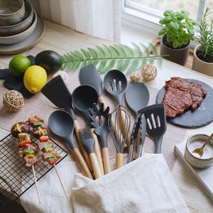 May include: A set of dark gray silicone kitchen utensils with light wooden handles, including a spatula, ladle, slotted spoon, pasta server, whisk, tongs, and basting brush, arranged together in a white fabric pouch on a wooden surface.
